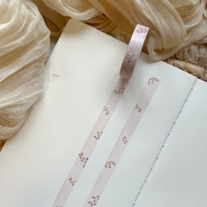 May include: Two rolls of pink washi tape with a delicate floral pattern. The tape is on a white notebook.