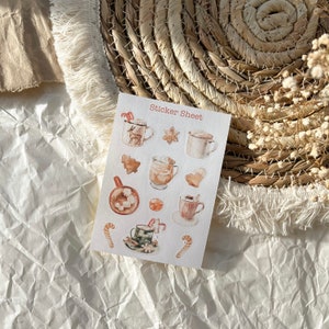 Könnte beinhalten: Ein Stickerbogen mit Aquarellillustrationen von festlichen Tassen, Lebkuchen und Zuckerstangen. Die Tassen haben verschiedene Designs, darunter ein Rentier. Der Text "Sticker Sheet" ist oben aufgedruckt.