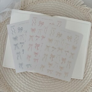 Könnte beinhalten: Drei Blätter Stickerpapier mit verschiedenen pastellfarbenen Schleifendesigns. Die Stickerblätter sind mit "Sticker Sheet" beschriftet.