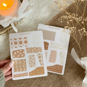 Könnte beinhalten: Zwei Blätter mit Aufklebern im zerrissenen Papier-Look. Die Aufkleber zeigen verschiedene Muster in Braun- und Beigetönen, darunter Sonnen, Monde, Schlangen und abstrakte Designs. Der Text "Sticker Sheet" ist auf jedem Blatt gedruckt.