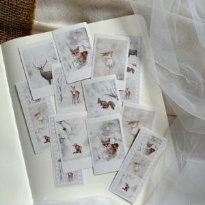 Könnte beinhalten: Ein Set mit 10 winterlichen, druckbaren Aufklebern mit verschiedenen Tieren, darunter ein Fuchs, ein Reh, ein Eichhörnchen und Vögel. Die Aufkleber sind im Aquarellstil mit weißem Hintergrund und dezenten grauen Akzenten gestaltet.