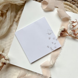 Può includere: Un blocco note adesivo quadrato bianco con un disegno a linea nera di una farfalla e piccoli fiori. Il blocco note è su una superficie bianca con un pezzo di tessuto beige e fiori secchi.