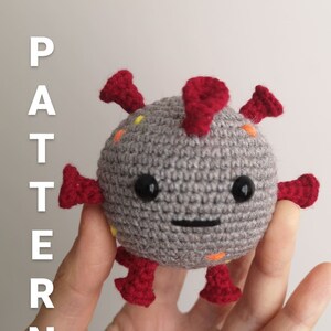 May include: A gray crocheted coronavirus plush toy with red spikes and orange and yellow dots. The toy has a smiling face with black eyes and a black mouth.