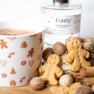 May include: A white mug with gingerbread cookie designs, filled with a brown liquid, next to a clear glass bottle with a label that reads "toasty gingerbread reed diffuser", vegan and handmade, surrounded by gingerbread cookies, cinnamon sticks, and nutmeg.
