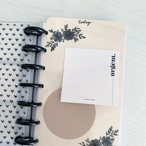 May include: A cream-colored planner page with black floral designs and the word "today" at the top. A white sticky note with the word "urgent" written vertically on it is attached to the page. A brown circle is in the center of the page.