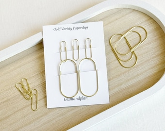 May include: Gold Variety Paperclips by Glamandplan. A set of gold paperclips in different shapes, including oval and rectangular, displayed on a wooden tray.