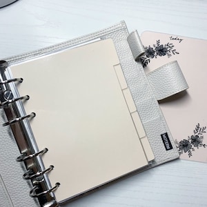 May include: A white leather bound planner with a clear plastic insert for a page. The planner has a silver ring binder and a black tab that says "MOTERM". The planner is open to a blank page. The planner is sitting on a white surface. A pink planner with the word "today" and a floral design is visible in the background.