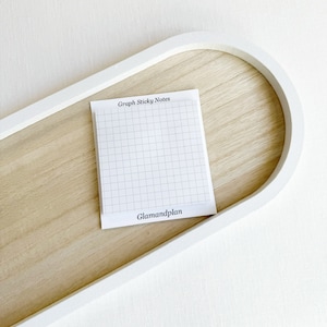 May include: A white graph sticky note pad with the text "Graph Sticky Notes" and "Glamandplan" on a light wood tray with a white border. The grid is light gray.