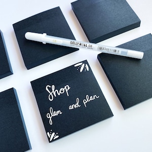May include: Black square sticky notes with white handwritten text that says "Shop glam and plan" on one of the notes. A white gel pen is laying on top of the notes.