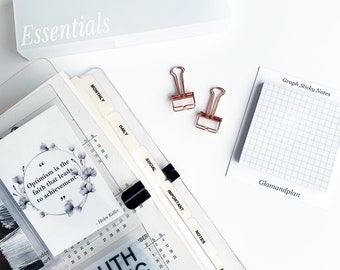 May include: A collection of stationery items, including a clear planner with labeled dividers, a white box labeled "Essentials", two rose gold binder clips, and a notepad with grid lines. The planner includes a quote by Helen Keller.