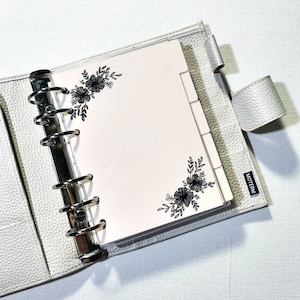 May include: A white leather planner with a clear plastic insert. The insert has a pink background with black floral designs in the corners. The planner has a silver ring binder and the word "MOTERM" is printed on the cover.
