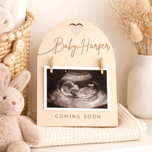 May include: A wooden pregnancy announcement sign with the name "Baby Harper" and a heart cutout. It displays a sonogram image held by small wooden clips. The text "COMING SOON" is printed below the sonogram.