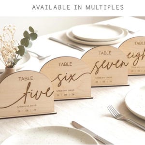 May include: Wooden table number signs with arched tops, displaying the numbers five, six, seven, and eight in elegant script. Each sign includes the names "Chloe and Jacob" and a date. The signs are set on a table with place settings.