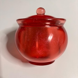 May include: A small, round, red glass jar with a matching lid. The jar has a spherical body and a small, round base. The lid is topped with a small, round knob. The jar appears to be made of a translucent material with glitter.