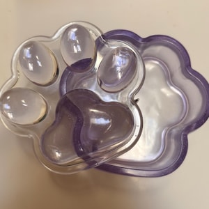 May include: A clear, paw-shaped container with a purple rim and lid. The lid features the paw print design, with rounded pads. The container is made of transparent plastic.