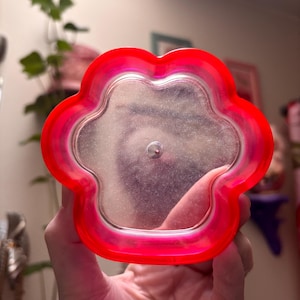 May include: A clear, flower-shaped container with a bright red rim. The container has a silver-colored interior and a small, silver-colored metal piece in the center. The container is held in a person's hand.