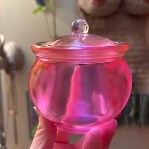 May include: A pink glass jar with a lid and a clear knob on top. The jar is round with a slightly flared rim. The jar is held in a hand.