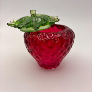 May include: A red glass strawberry-shaped jar with a green glass lid. The jar has a textured surface and the lid has a leaf design.