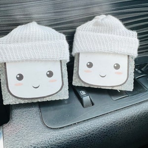 May include: Two white car air fresheners shaped like a person wearing a white knit hat. Each air freshener has a smiling face with rosy cheeks and black eyes. The air fresheners are sitting on the dashboard of a car.