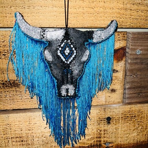 May include: A black and white bull skull with blue fringe hanging from a wooden wall. The skull has a diamond pattern on the forehead and is decorated with white and blue beads.