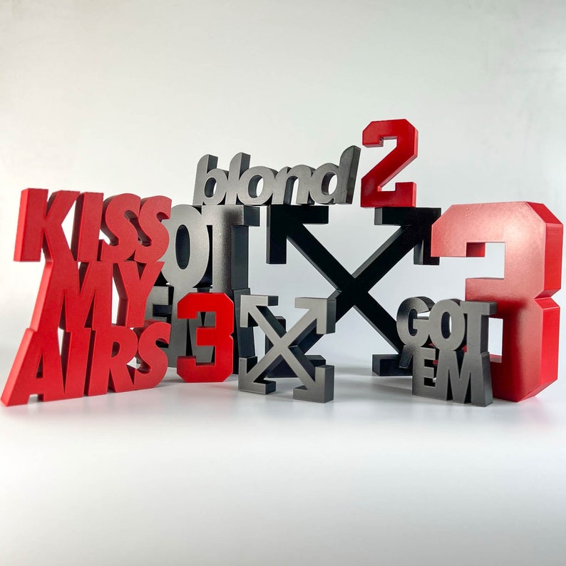 Peut inclure: Un ensemble de lettres 3D noires et rouges &eacute;pellant "Kiss My Airs Got Em" et "Blond 2" avec un X noir et blanc au milieu.