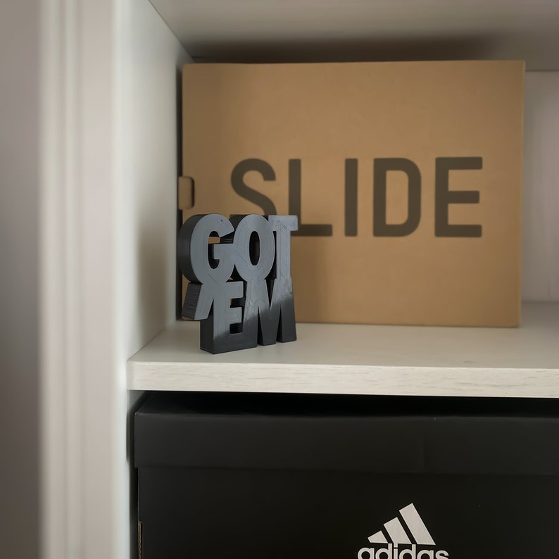 Peut inclure: Lettres en bois noir en forme de blocs orthographiant "GOT 'EM" sur une &eacute;tag&egrave;re blanche avec une bo&icirc;te brune derri&egrave;re qui dit "SLIDE" en lettres noires. L'&eacute;tag&egrave;re est au-dessus d'une bo&icirc;te noire avec le logo Adidas.