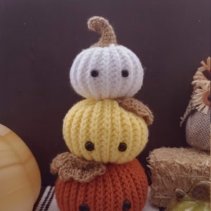 Puede incluir: Una pila de tres calabazas de ganchillo, con una calabaza blanca en la parte superior, una calabaza amarilla en el medio y una calabaza naranja en la parte inferior. Cada calabaza tiene ojos de botón negros.