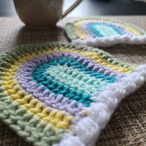 Könnte beinhalten: Zwei handgefertigte Regenbogen-Untersetzer aus Häkelarbeit. Jeder Untersetzer hat ein halbkreisförmiges Design mit Bändern in Grün, Gelb, Lila und Türkis. Ein weißer, wolkenartiger Rand umrahmt die Unterkante. Eine Tasse ist im Hintergrund.