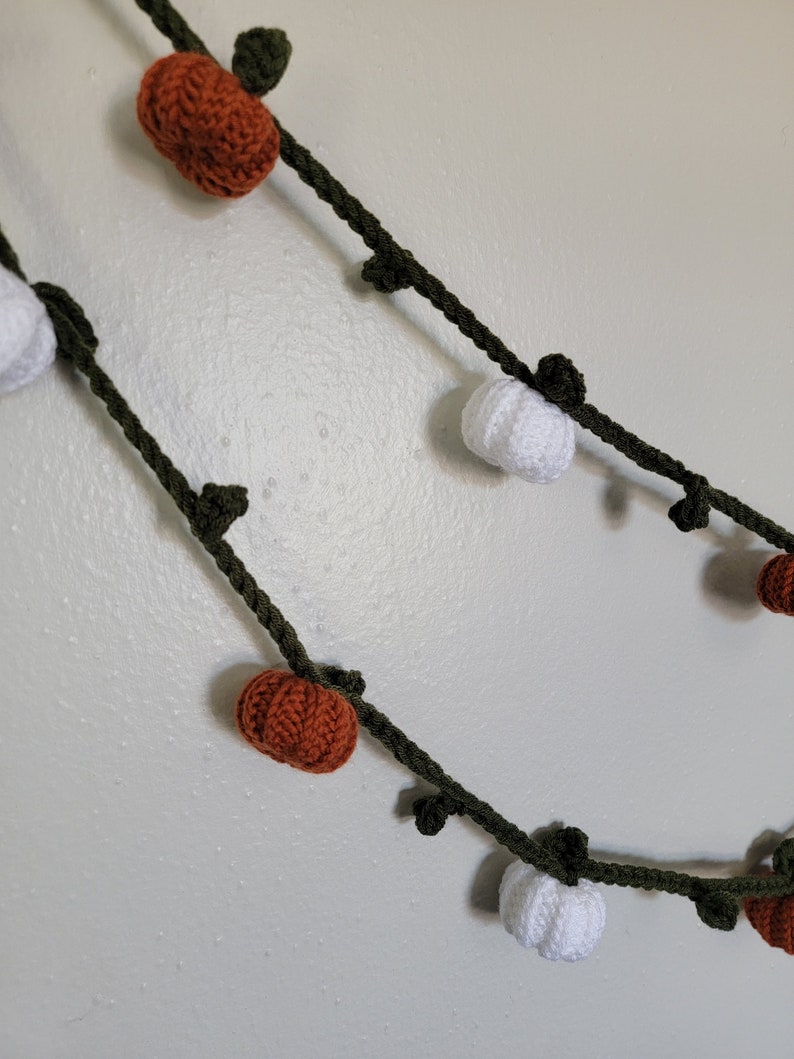 May include: A green garland with crocheted orange and white pumpkins. The garland is perfect for fall decor.