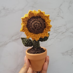 Op de afbeelding: Een gehaakte zonnebloem in een terracotta pot. De zonnebloem is geel en bruin, met groene bladeren. De pot is gevuld met bruine aarde.