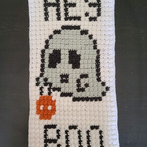 May include: White crocheted wall hanging with black and grey yarn depicting a ghost with a skull face and the words "Hey Boo".