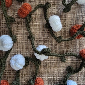 May include: A crocheted garland with alternating white and orange pumpkins. The garland is made of green yarn and is perfect for fall decor.