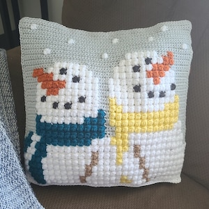 Puede incluir: Una almohada tejida a crochet con un tema invernal que presenta dos muñecos de nieve. Los muñecos de nieve están hechos con hilo blanco y tienen ojos negros, narices naranjas y bufandas de color turquesa. El fondo es gris claro con puntos blancos.