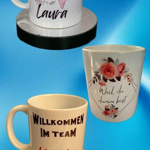 Könnte beinhalten: Weiße Keramikbecher mit personalisiertem Text und floralen Designs. Ein Becher trägt die Aufschrift "Willkommen Laura" mit einem floralen Initial. Ein anderer sagt "Willkommen im Team Kirsten". Der dritte Becher hat den Text "Weil du darum bist".
