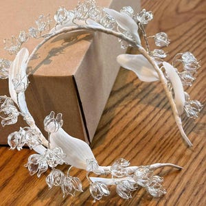 May include: A headband featuring a series of clear, translucent flower-shaped beads attached to a silver-toned wire frame. The structure is accented with smooth, white fabric leaves wrapped around the band, creating a floral arrangement.