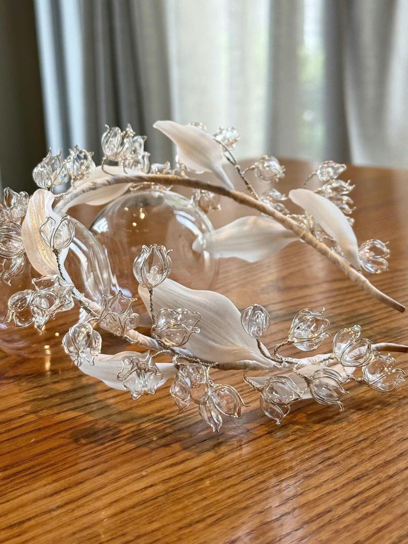 May include: A headband featuring a series of clear, bell-shaped glass or crystal flowers and smooth, opaque white leaves attached to a thin, light-colored band. The floral elements are connected by thin, metallic wire stems.