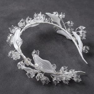 Peut inclure: Un serre-t&ecirc;te orn&eacute; d'une s&eacute;rie de fleurs en verre transparent en forme de cloche et de feuilles blanches opaques, lisses et allong&eacute;es. Les &eacute;l&eacute;ments floraux sont fix&eacute;s sur une fine armature m&eacute;tallique flexible recouverte de tissu blanc.