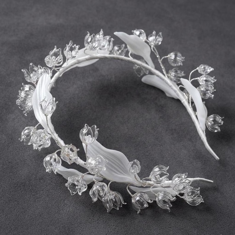 Peut inclure: Un serre-t&ecirc;te orn&eacute; d'une s&eacute;rie de fleurs en verre transparent en forme de cloche et de feuilles blanches opaques, lisses et allong&eacute;es. Les &eacute;l&eacute;ments floraux sont fix&eacute;s sur une fine armature m&eacute;tallique flexible recouverte de tissu blanc.