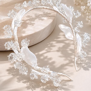 Peut inclure: Un serre-t&ecirc;te argent&eacute; orn&eacute; de d&eacute;licates fleurs en cristal transparent rappelant le muguet et de feuilles en tissu blanc lisse. Les &eacute;l&eacute;ments floraux sont fix&eacute;s sur une fine armature m&eacute;tallique flexible recouverte d'un ruban blanc.