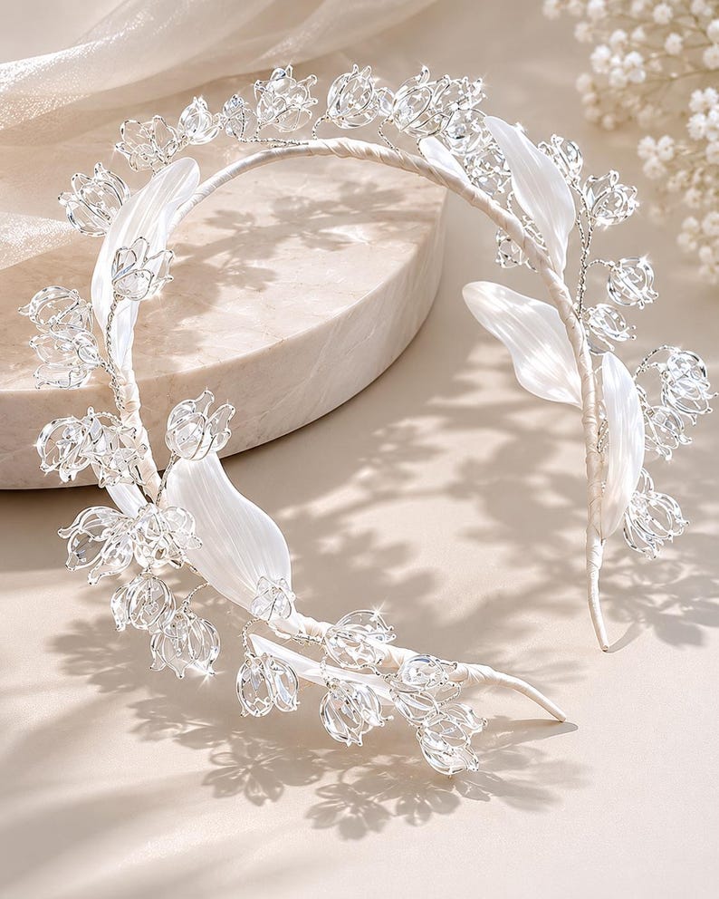 Peut inclure: Un serre-t&ecirc;te argent&eacute; orn&eacute; de d&eacute;licates fleurs en cristal transparent rappelant le muguet et de feuilles en tissu blanc lisse. Les &eacute;l&eacute;ments floraux sont fix&eacute;s sur une fine armature m&eacute;tallique flexible recouverte d'un ruban blanc.
