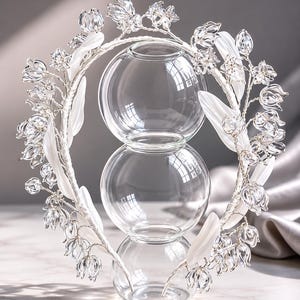 Peut inclure: Un serre-t&ecirc;te circulaire orn&eacute; de fleurs transparentes en forme de cloche et de feuilles blanches lisses fix&eacute;es sur une armature envelopp&eacute;e. Le serre-t&ecirc;te entoure un vase en verre transparent compos&eacute; de trois sph&egrave;res empil&eacute;es de tailles vari&eacute;es.