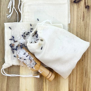 May include: Several small, off-white muslin bags with drawstring closures are arranged on a wooden surface. One bag is open, revealing lavender buds and salt, with a small wooden scoop. The bags are likely for aromatherapy or potpourri.