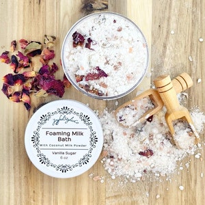 Puede incluir: Un recipiente blanco de baño de leche espumosa con azúcar de vainilla, 170 g, y un recipiente transparente de sales de baño con pétalos de rosa. Dos pequeñas cucharas de madera y pétalos de rosa dispersos sobre una superficie de madera.