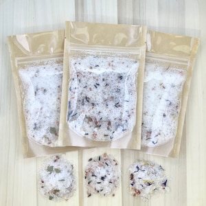 Puede incluir: Tres bolsas transparentes llenas de sales de baño blancas y botánicos secos. Las bolsas tienen un respaldo de papel marrón. Debajo, tres pequeños montones de sales de baño con diferentes mezclas botánicas.