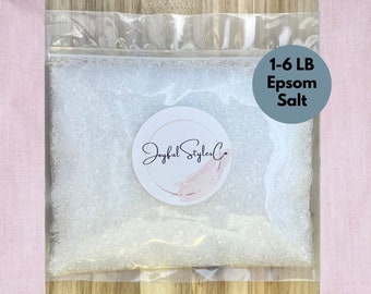 May include: A clear plastic bag filled with white Epsom salt. The bag has a circular sticker with the text "Joyful Styler Co" and a blue circle with the text "1-6 LB Epsom Salt". The background is a light pink fabric.