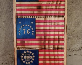 May include: Wooden American flag art piece with a distressed finish. The flag design features red and white stripes, a blue field with stars, and the number "76" in the center. The bottom section includes the text "1776 250 2026".