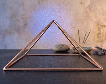 May include: A copper-colored metal pyramid frame with a square base. The structure is composed of thin, smooth rods connected at a single apex point, creating a geometric open-frame design that sits on a flat surface.