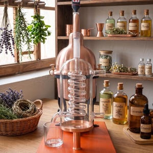 May include: A copper and glass distillation apparatus featuring a central glass condenser coil, a copper boiler base, and a digital thermometer inserted at the top. The unit is mounted on a rectangular wooden board with a small glass collection cup.