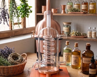 May include: A copper and glass distillation apparatus featuring a central glass condenser coil, a copper boiler base, and a digital thermometer inserted at the top. The unit is mounted on a rectangular wooden board with a small glass collection cup.