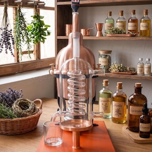 Op de afbeelding: Een destilleerapparaat van koper en glas met een centrale glazen condensor-spiraal, een koperen ketelbasis en een digitale thermometer aan de bovenkant. Het apparaat is gemonteerd op een rechthoekige houten plank met een klein glazen opvangbakje.